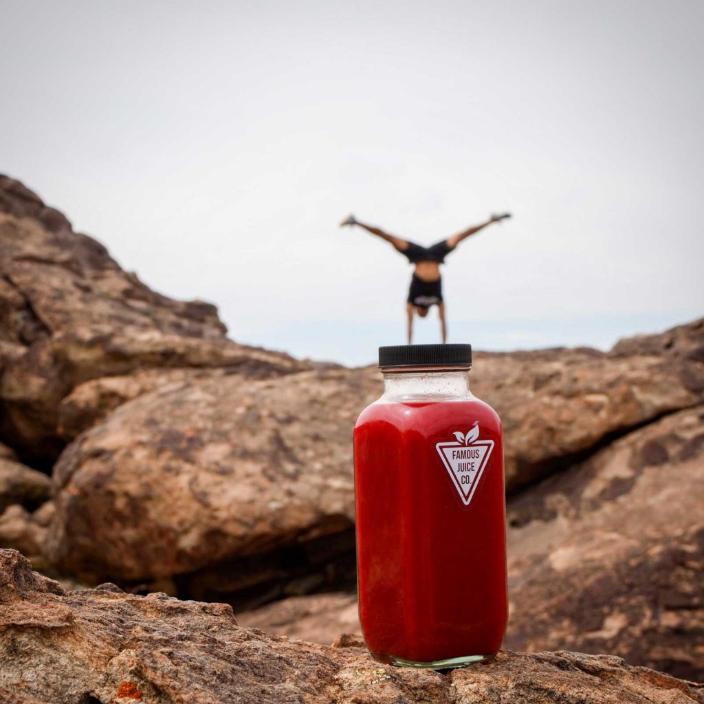 Image of a cold pressed juice bottle in the foreground with a person doing a hand stand in the background